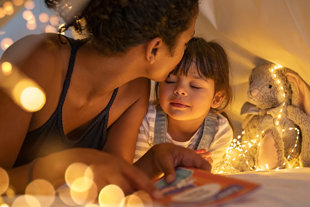 A mother kissing her young daughter on the forehead inside a fairy-lit reading tent at bedtime