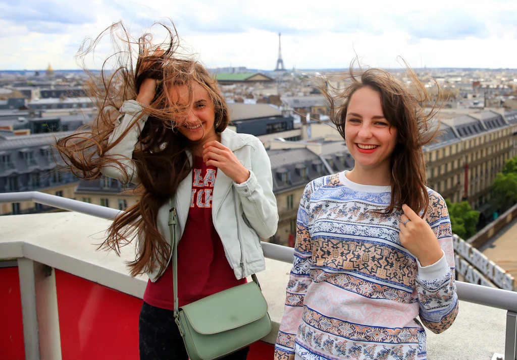Two friends laughing on a Paris rooftop with the Eiffel Tower in the distance, hair caught in the wind